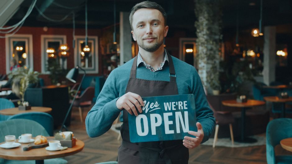 Man in apron holding a “Yes, We’re Open” sign inside a cozy, modern café with tables, chairs, and food on the tables in the background.